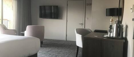 Hotellihuone, jossa on sänky, kaksi tuolia, työpöytä, kahvinkeitin, televisio ja ovi.