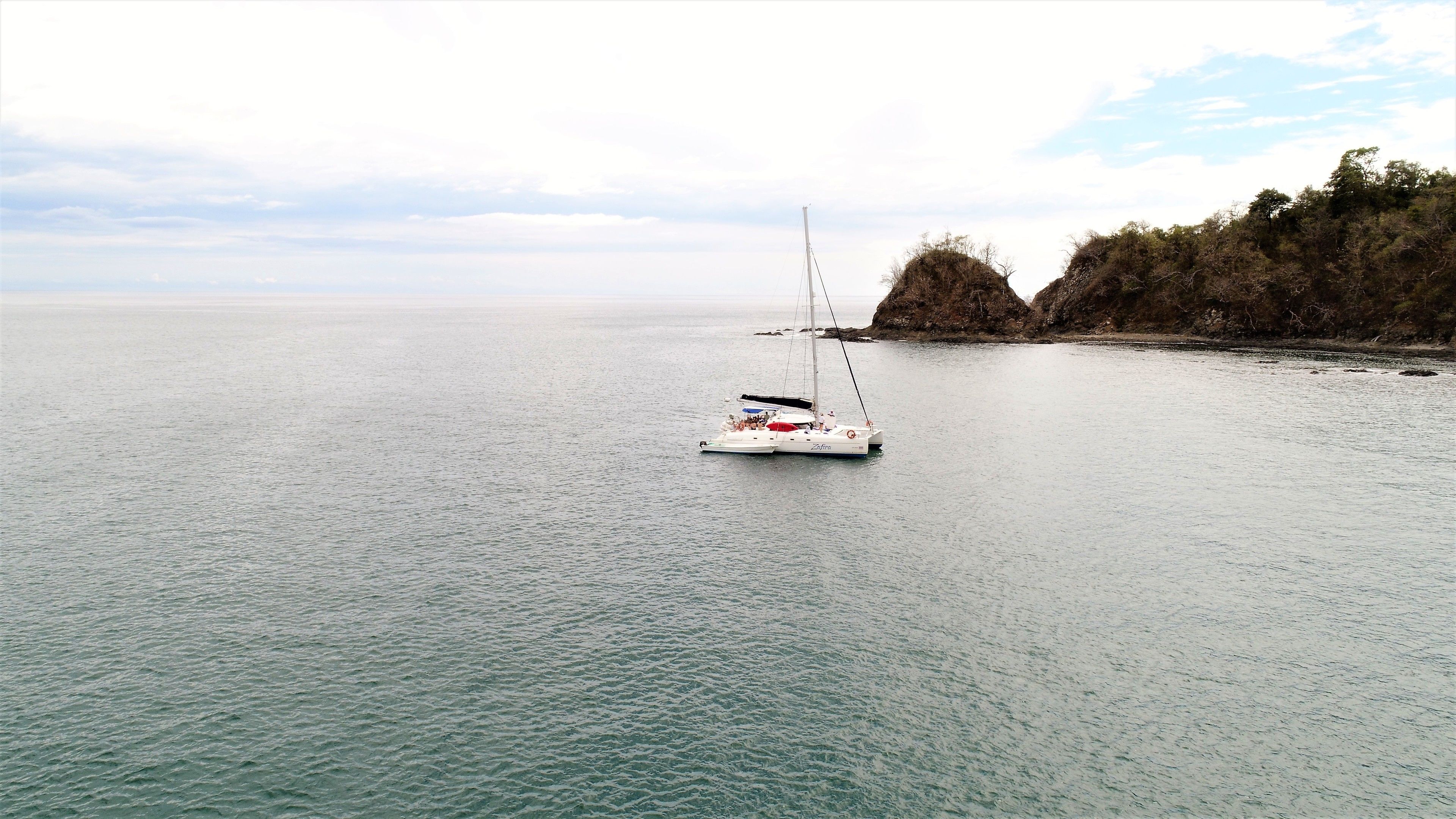 Un velero en aguas tranquilas, con una costa rocosa y vegetación escasa al fondo.