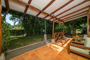 A covered outdoor area with a wooden bench and a table, surrounded by greenery and a tiled floor. - Hotel Palma de Oro (Bejuco)