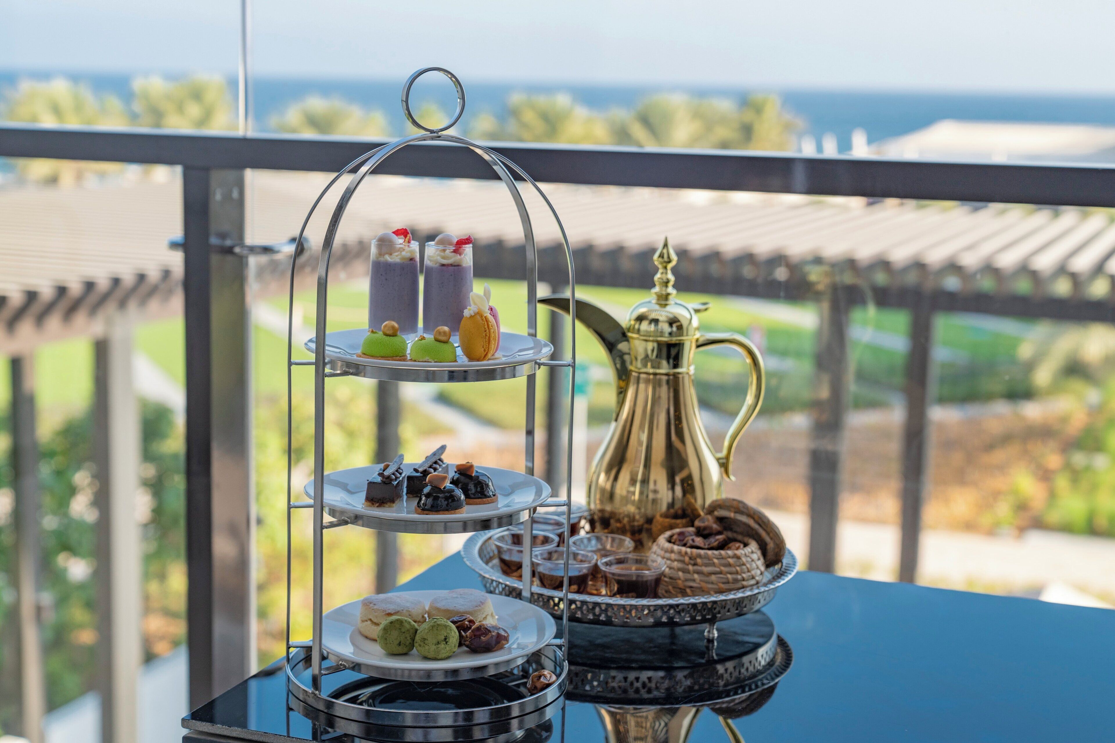 A tiered dessert stand with various sweets and a decorative pitcher on a table with a view of the outdoors.