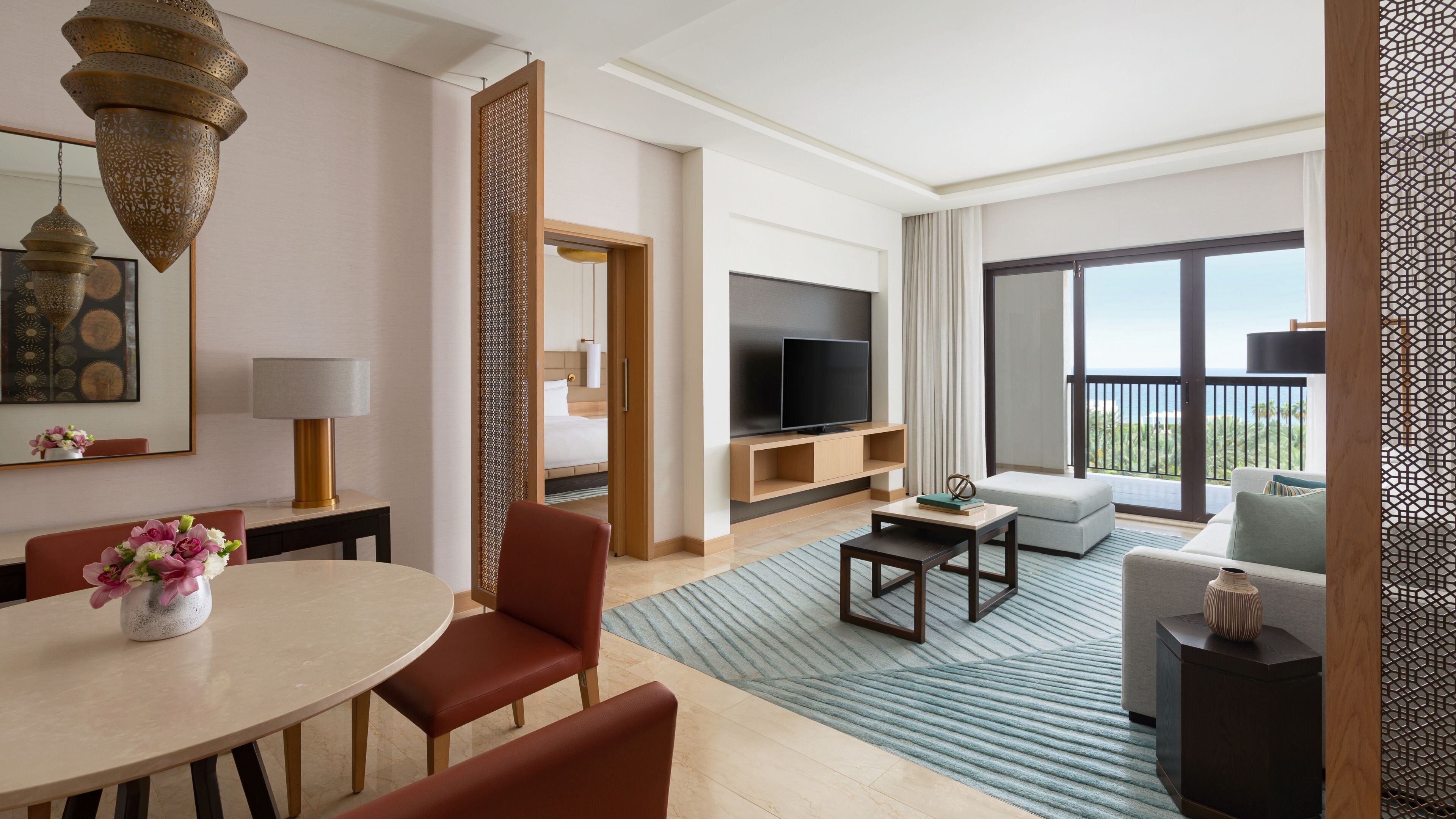 A modern hotel room with a dining area, a sofa, a TV, and a balcony with a view.
