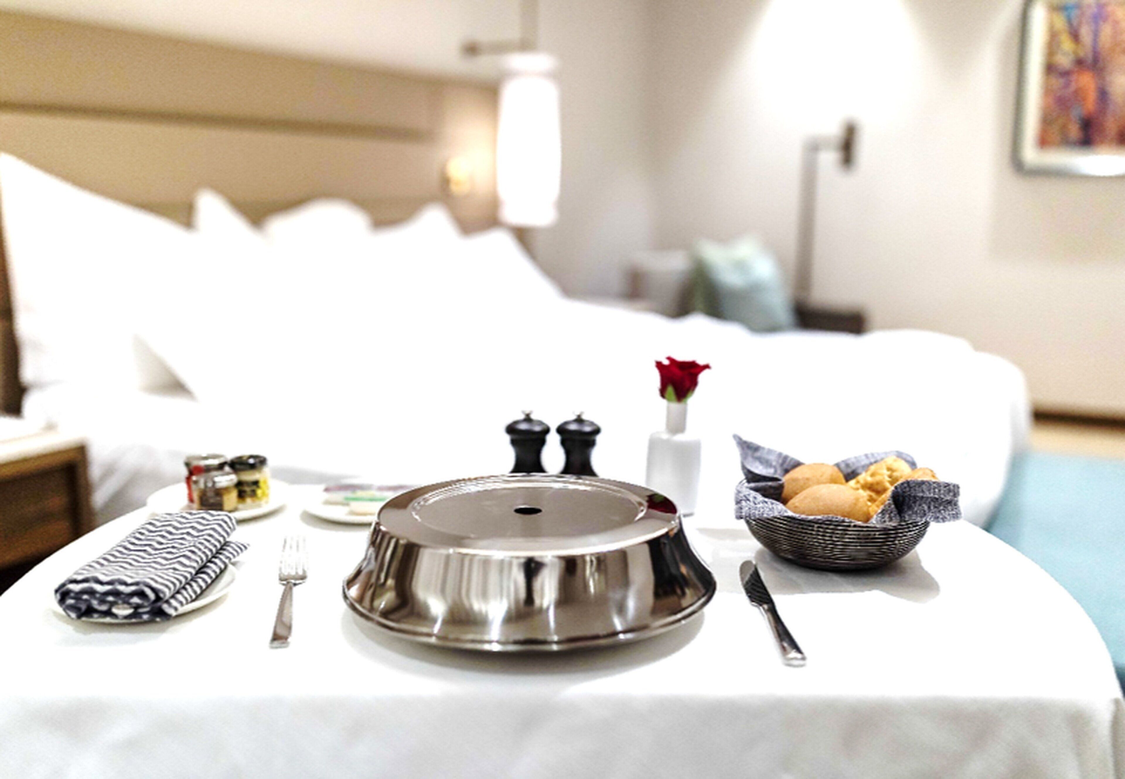 Una mesa de desayuno ordenada con una bandeja de metal, un bol de pan, una rosa, saleros y pimenteros, un tenedor y una servilleta.