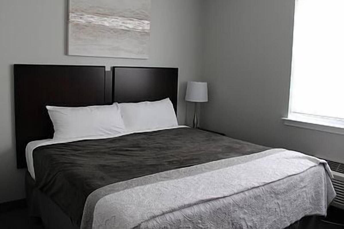 A neatly made bed with a dark headboard, a bedside lamp, and a window with blinds.