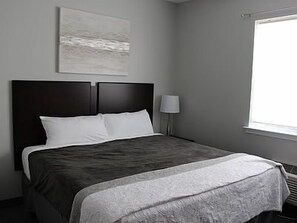 Una cama bien hecha con un cabecero oscuro, una lámpara de noche y una ventana con persianas.