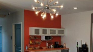 Uma sala de estar moderna com um sofá, área de cozinha com armários brancos, micro-ondas e um lustre singular.
