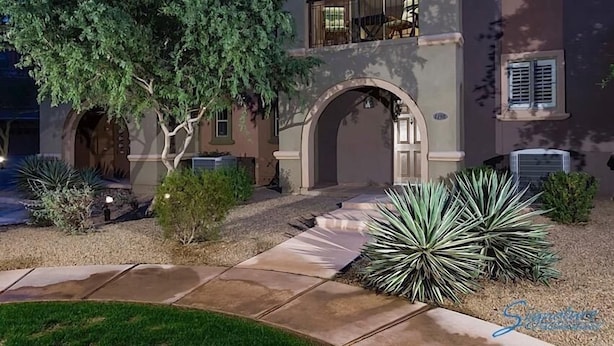 A building with a stone pathway leading to an arched entrance, surrounded by various plants and a tree. - Hot Spot at Desert Ridge By Signature Vacation R (Phoenix)