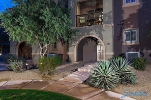 A building with a stone pathway leading to an arched entrance, surrounded by various plants and a tree.