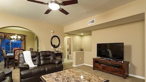 A living room with a ceiling fan, a leather sofa, a TV on a wooden stand, and a dining area with a chandelier. - Hot Spot at Desert Ridge By Signature Vacation R (Phoenix)