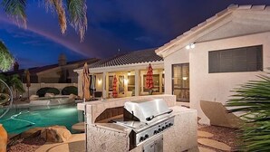 An outdoor pool area with a stone barbecue grill, lounge chairs, and umbrellas. - Hole in One By Signature Vacation Rentals (Goodyear)
