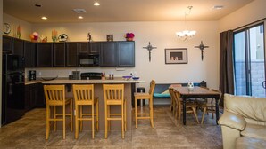 A modern kitchen with dark cabinets, a central island with bar stools, a dining area with a table and chairs, and a large window with curtains. - Heart of Tucson By Signature Vacation Rentals (Tucson)