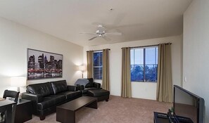 Un salon moderne avec un canapé en cuir noir, une table basse, un téléviseur et un ventilateur de plafond. Il y a deux fenêtres avec des rideaux, et un tableau représentant un paysage urbain est accroché au mur.