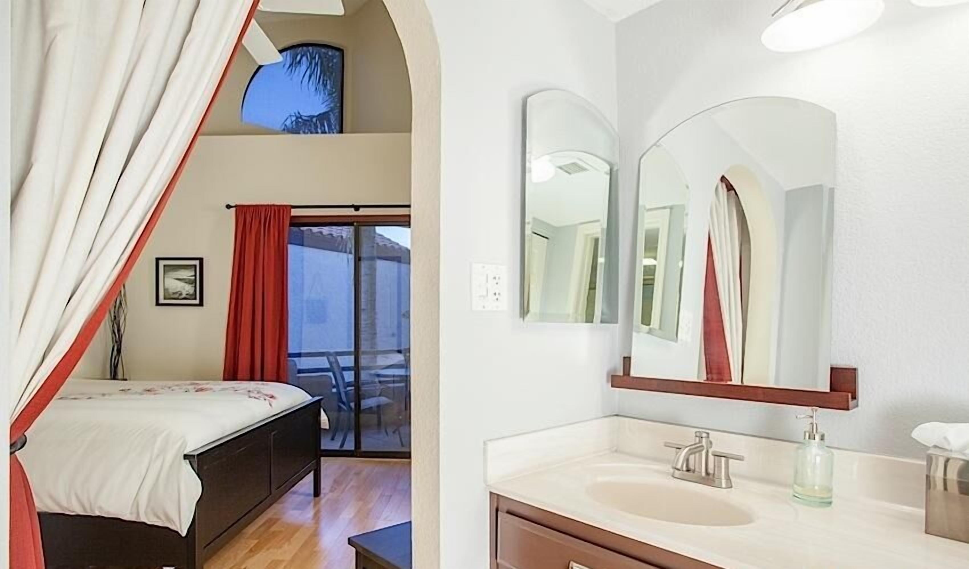 Une chambre à coucher avec un lit, une fenêtre avec des rideaux et un balcon avec une chaise. La salle de bain est équipée d’un lavabo, d’un miroir et d’un distributeur de serviettes.