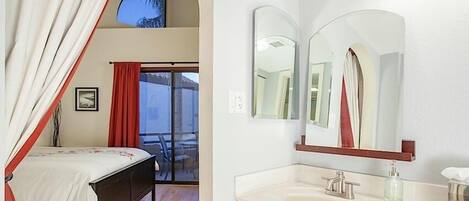 Ein Schlafzimmer mit Bett, einem Fenster mit Vorhängen und einem Balkon mit Stuhl. Das Badezimmer verfügt über ein Waschbecken, einen Spiegel und einen Handtuchspender.
