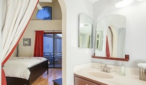 A bedroom with a bed, a window with curtains, and a balcony with a chair. The bathroom has a sink, a mirror, and a towel dispenser. - Happy Ours By Signature Vacation Rentals (Paradise Valley)
