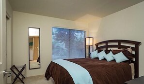 A bedroom with a bed, a mirror, a window with blinds, and a nightstand with a lamp.