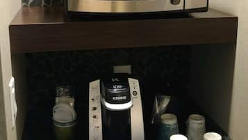 Una kitchenette con un microondas sobre una encimera oscura, una cafetera debajo y una variedad de cápsulas de café y tazas.