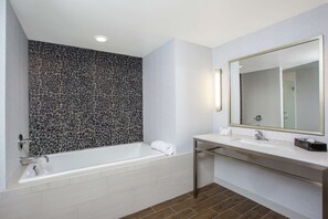 A modern bathroom with a stone mosaic wall, a white bathtub, a large mirror, and a white sink with a chrome faucet. - Hilton Garden Inn Olympia (Olympia)