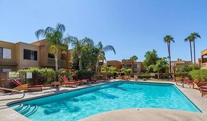 A swimming pool surrounded by palm trees and lounge chairs, with modern buildings in the background.