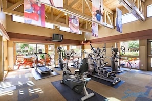 A gym with treadmills, elliptical machines, and stationary bikes. There are large windows and a TV on the wall.