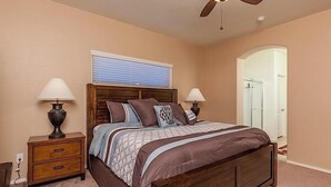 Un dormitorio con una cama de madera, dos mesitas de noche con lámparas, un ventilador de techo, una ventana con persianas y una puerta que da al baño.