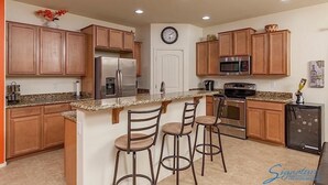 Una cocina con armarios de madera, un refrigerador de acero inoxidable, un microondas, una estufa y un lavamanos. Hay tres taburetes de bar y un reloj en la pared.
