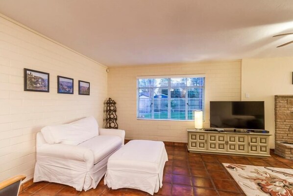 A living room with a white couch, a TV on a cabinet, a window with blinds, and a fireplace. - Encanto Bungalow By Signature Vacation Rentals (Phoenix)