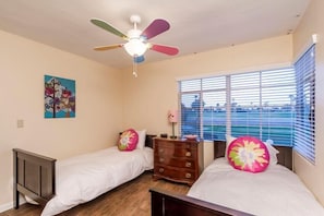 Une chambre avec deux lits simples, un ventilateur de plafond, une commode et une fenêtre avec des stores.