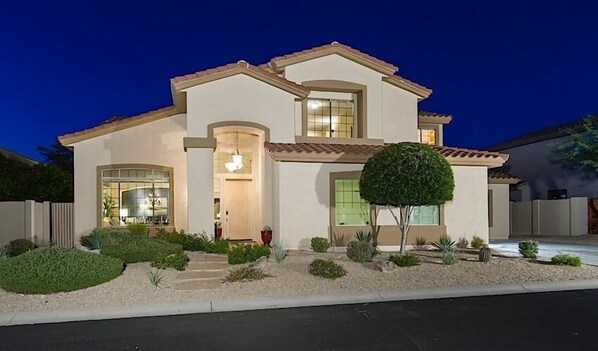 A two-story house with a light-colored exterior, a tiled roof, and a well-maintained front yard with various plants and a small tree. - Dynamite By Signature Vacation Rentals (Cave Creek)