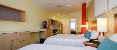 Habitación de hotel con dos camas, un escritorio, una silla y una pequeña cocina.