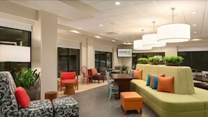A modern lobby with a green sofa, orange ottoman, and patterned chairs. There is a dining area with a table and chairs, and a sign that reads "HOME2". - Home2 Suites by Hilton Portland (Portland)
