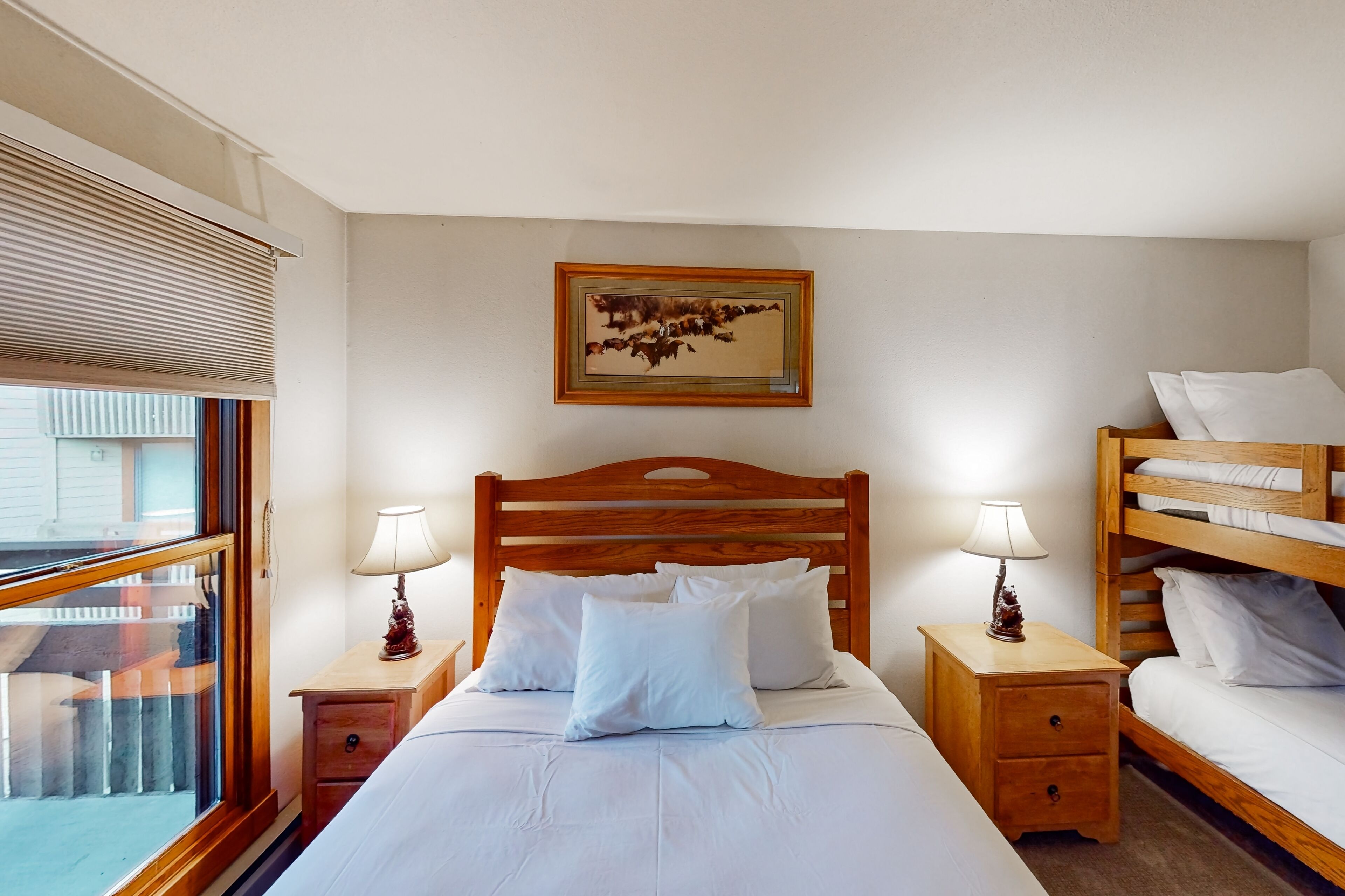 A bedroom with a wooden bed, a nightstand with a lamp, a bunk bed, and a window with blinds.