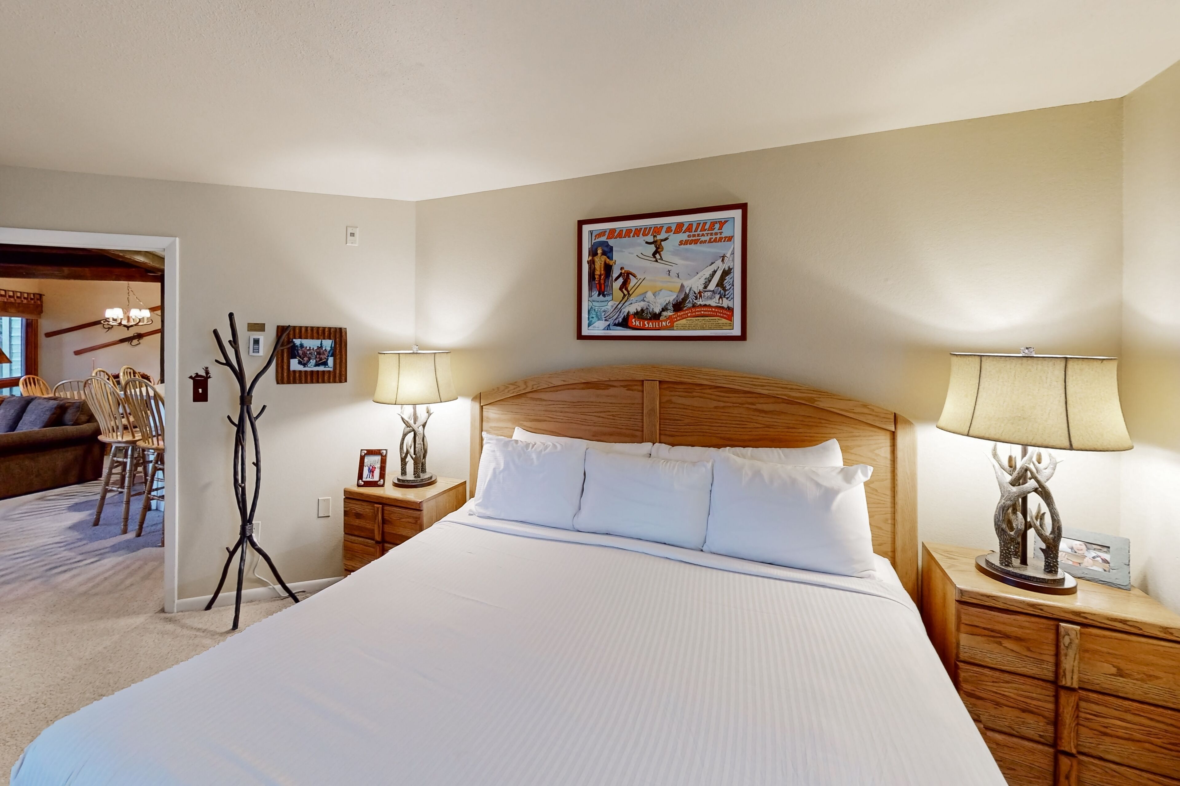 A hotel room with a large bed, wooden headboard, two bedside tables with lamps, a framed poster on the wall, and a wooden chair in the corner.