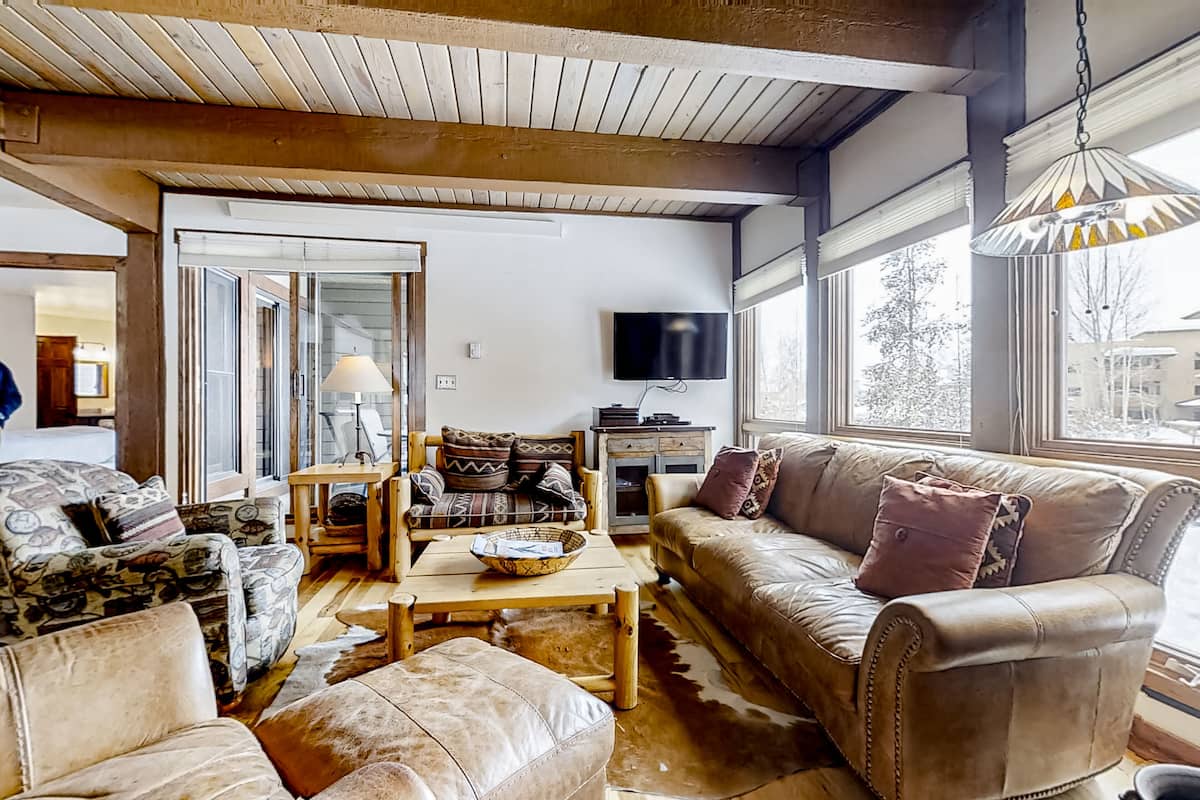 Ein gemütliches Wohnzimmer mit Sofa, Sessel und Couchtisch. Ein Fernseher ist an der Wand befestigt und ein Fenster sorgt für natürliches Licht.