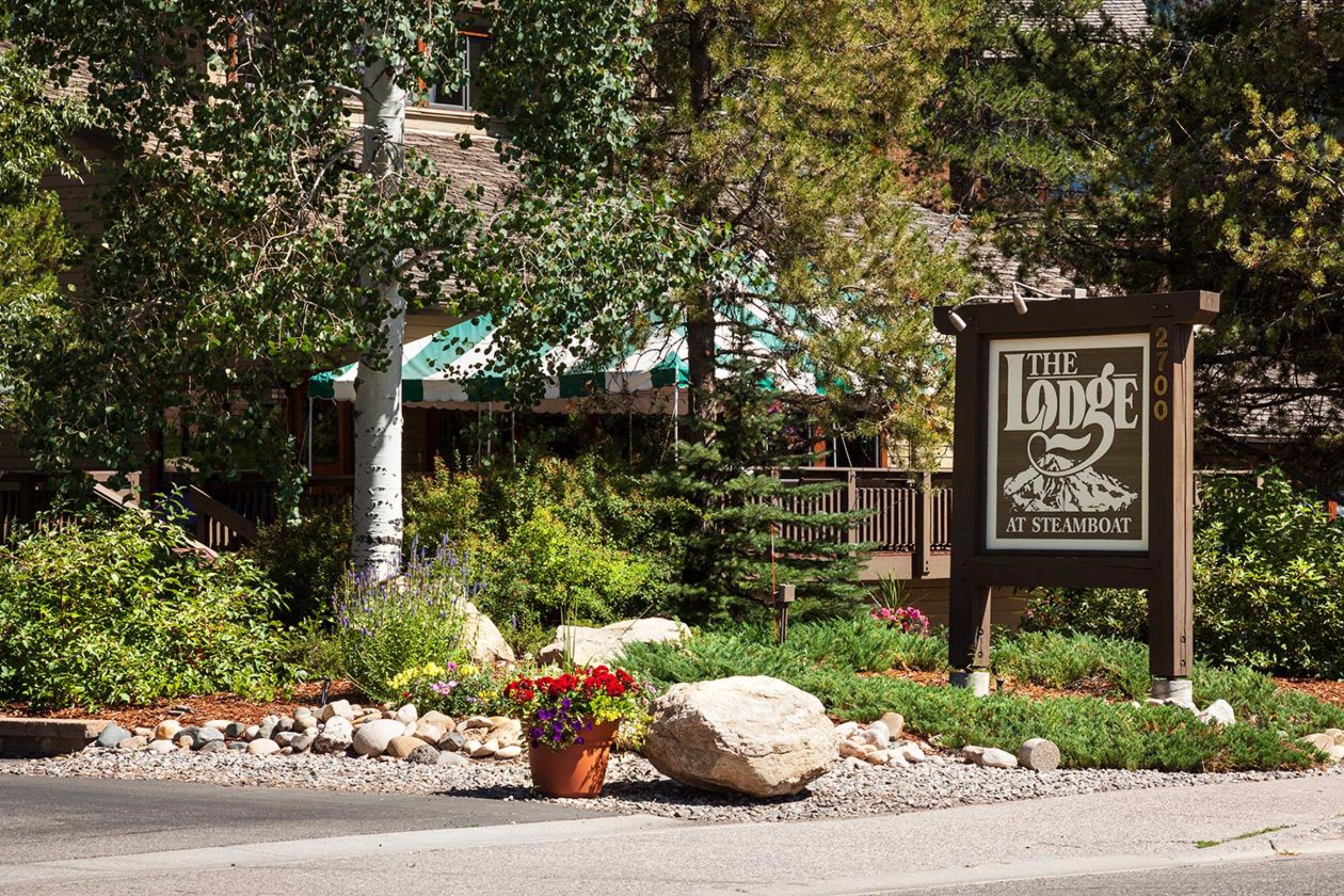 Señal de 'The Lodge at Steamboat' con un gráfico de montaña y tienda de campaña, rodeada de un jardín ajardinado con rocas y plantas en macetas.