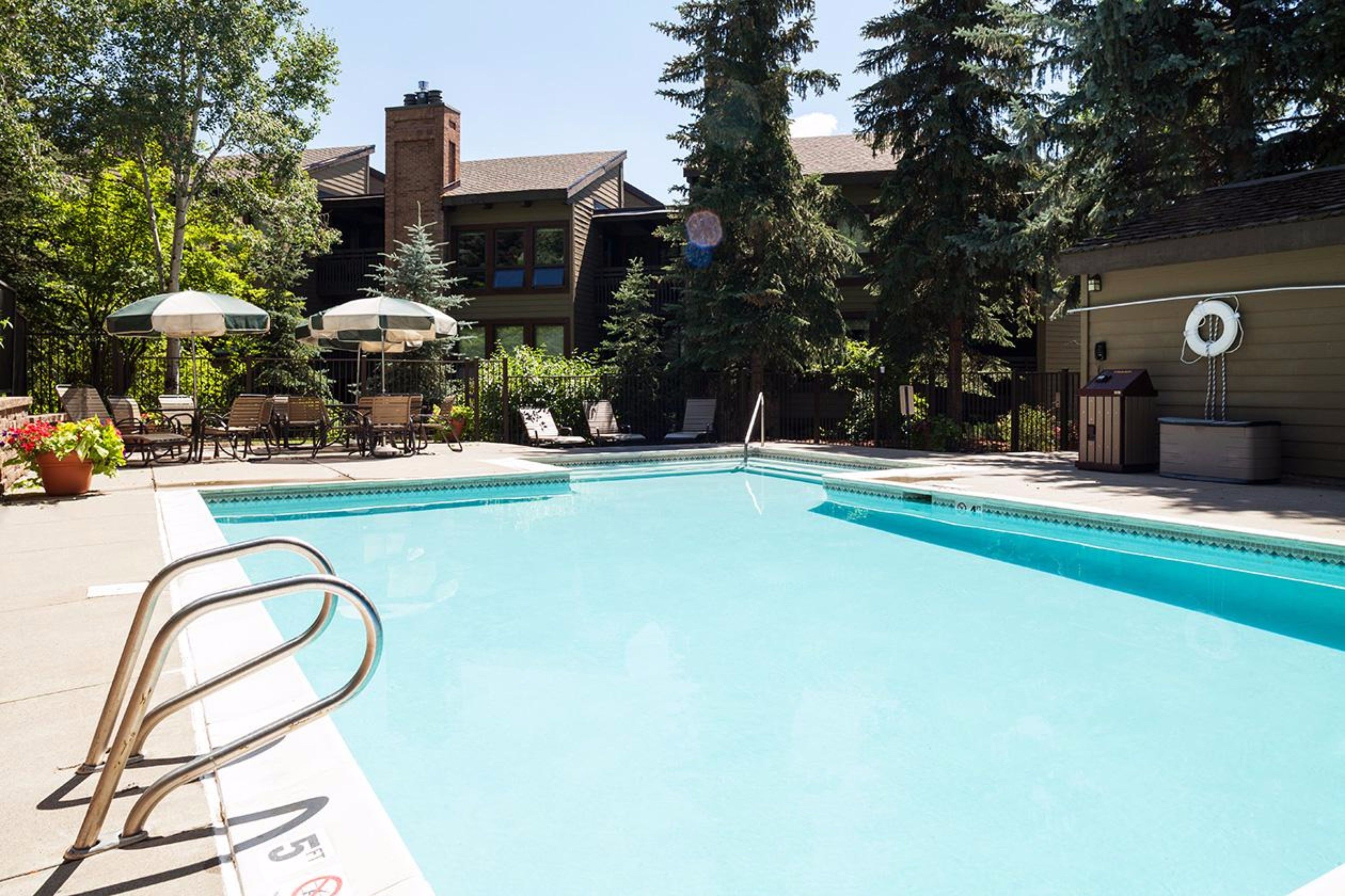 Une piscine entourée d’arbres, avec des chaises longues et des parasols, et un bâtiment doté d’une cheminée en briques.