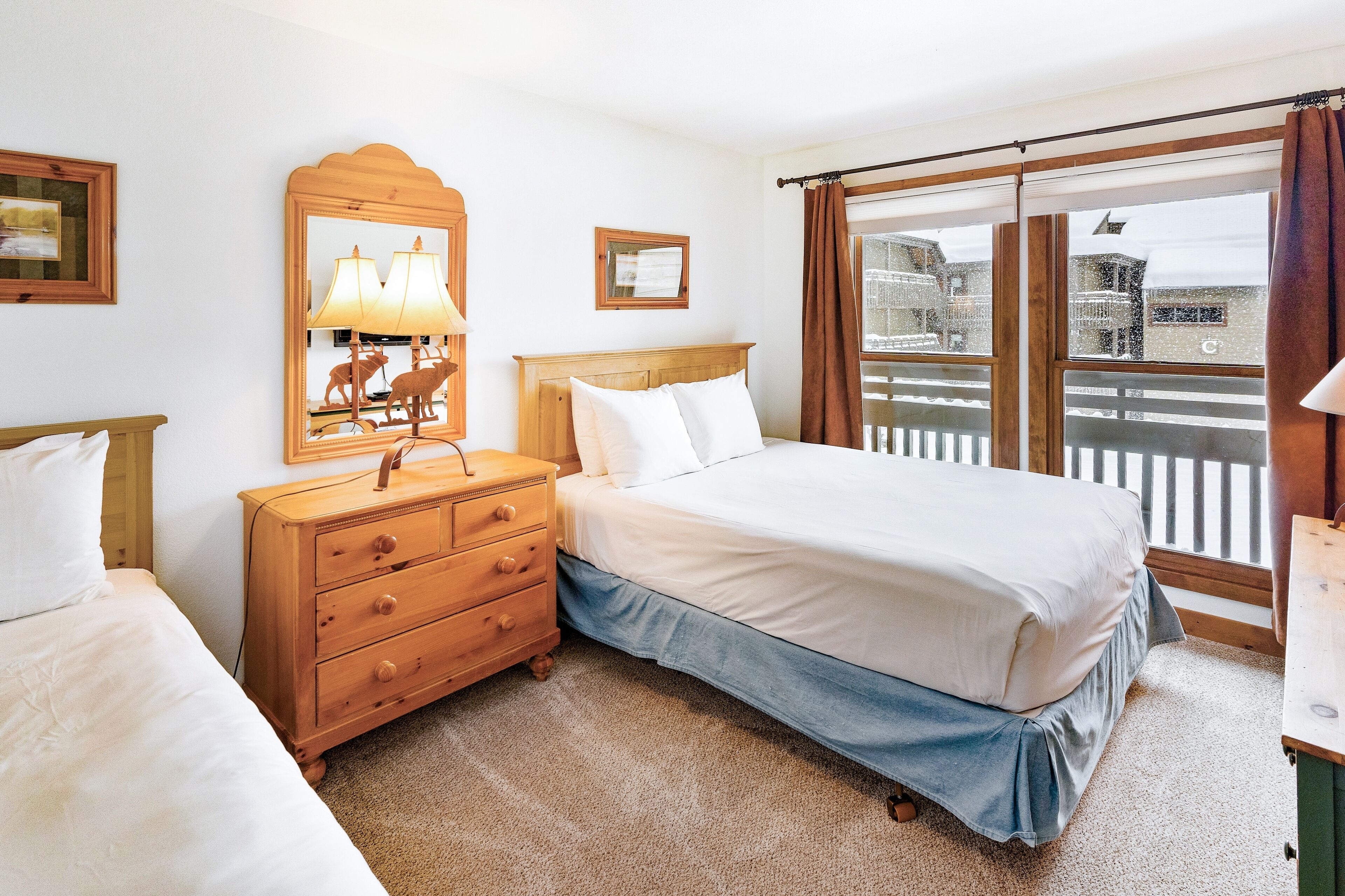 Un dormitorio con cama, cómoda de madera, espejo y ventana con vista a edificios.