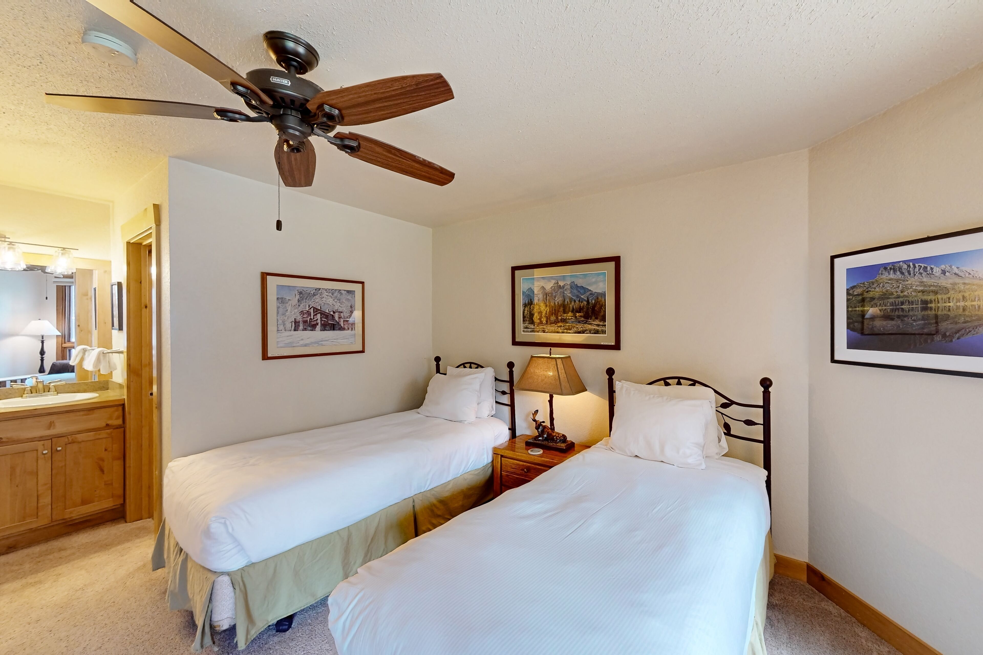 Une chambre d’hôtel avec deux lits simples, un ventilateur de plafond, une tête de lit en bois, une table de chevet avec une lampe et des tableaux encadrés accrochés aux murs.