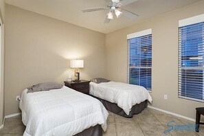 Un dormitorio con dos camas, un ventilador de techo, una mesita de noche con lámpara y ventanas con persianas.