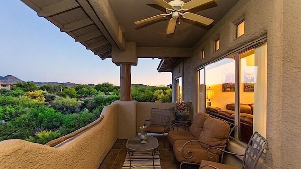 A balcony with a ceiling fan, a small table with a vase, and a sofa. The balcony overlooks a mountainous landscape with greenery.