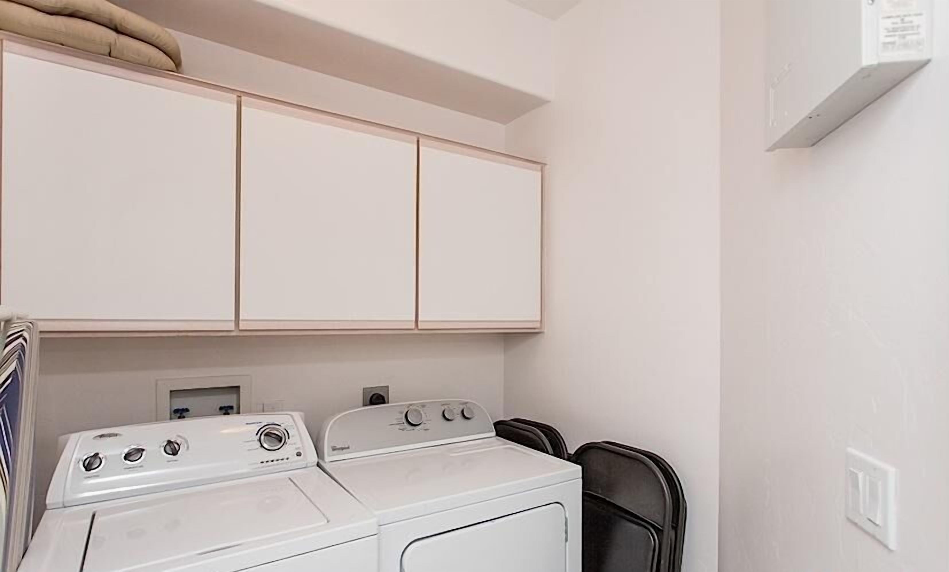 A laundry room with two washing machines and a dryer, white cabinets above, and a power outlet on the wall.