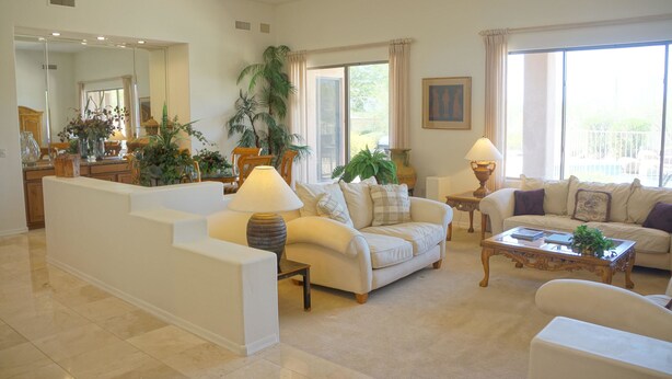 A spacious living room with a large sofa, a coffee table, and a dining area with a table and chairs. There are several potted plants and a large window providing natural light. - Troon Skyline Private By Signature Vacation Rental (Scottsdale)