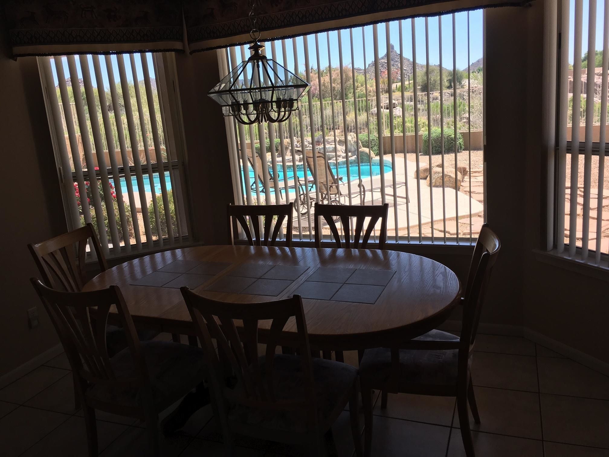 A dining area with a round table and chairs, large windows with vertical blinds, and a view of a swimming pool and mountains.