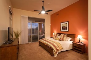 A bedroom with a bed, a ceiling fan, a nightstand, a lamp, a TV, and a sliding glass door leading to a balcony.