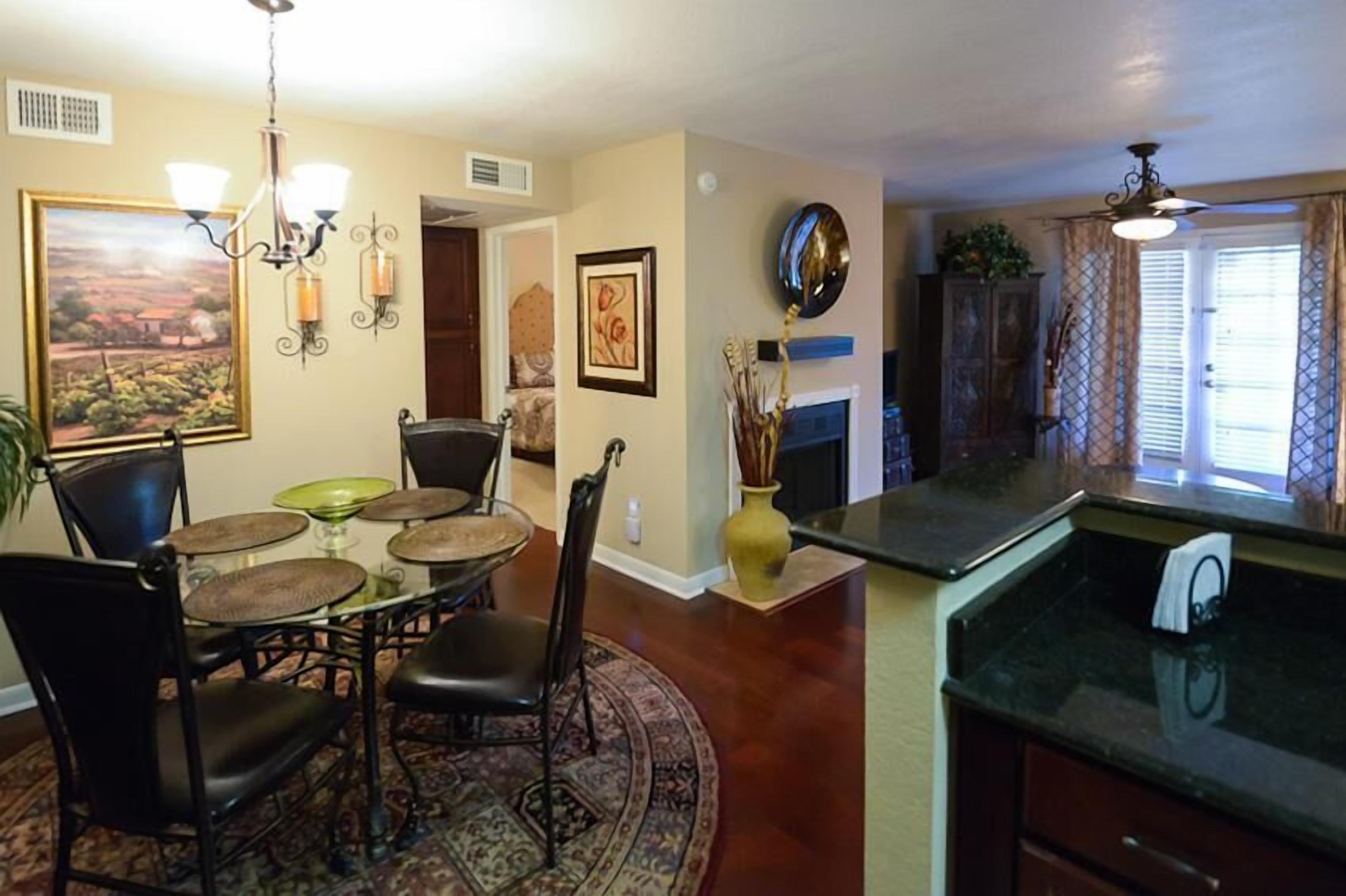 A dining area with a round table, chairs, and a chandelier. There is a fireplace with a vase of dried plants. The room has a wooden floor and beige walls.