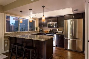 Une cuisine moderne dotée de plans de travail en granit, d’appareils électroménagers en acier inoxydable et de meubles de rangement sombres. On y trouve deux tabourets de bar et des luminaires suspendus au-dessus de l’îlot central.