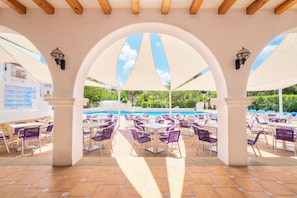 Un patio exterior con mesas y sillas, una piscina y un área cubierta con arcos.