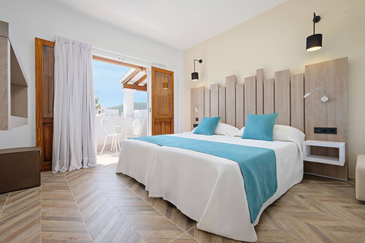 Une chambre avec un grand lit, une tête de lit en bois, une table de chevet blanche et une vue sur l’extérieur.
