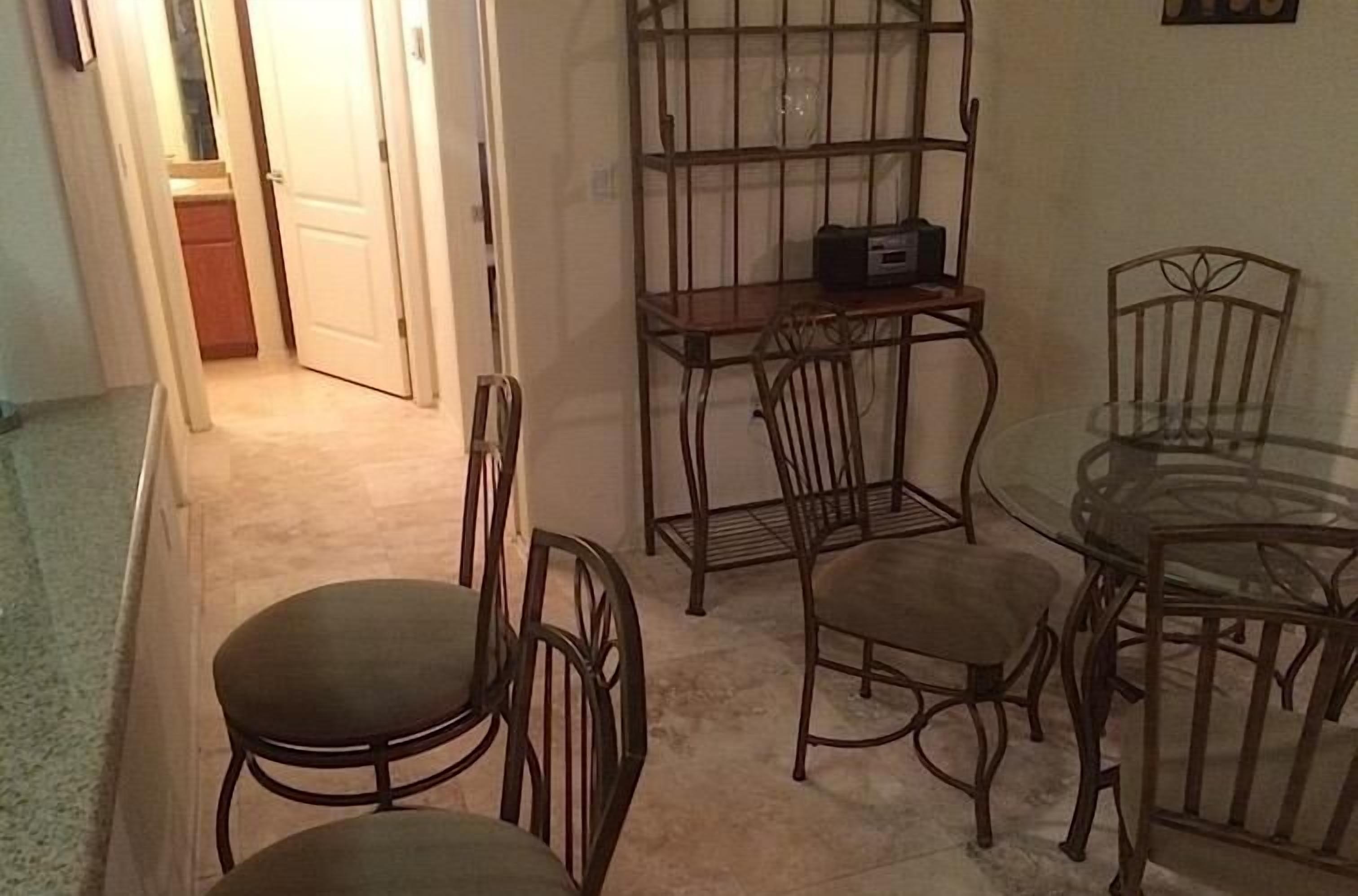 A dining area with a glass table and metal chairs, a small metal shelf with a radio, and a door leading to another room.
