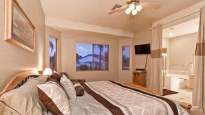 A bedroom with a large bed, a ceiling fan, a TV, and a bathroom visible through an open door. - Augusta On The Green by Signature Vacation Rentals (Mesa)