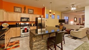 A modern kitchen with granite countertops, a central island, and stainless steel appliances. There is a dining area with a white sofa and a TV. - Augusta On The Green by Signature Vacation Rentals (Mesa)